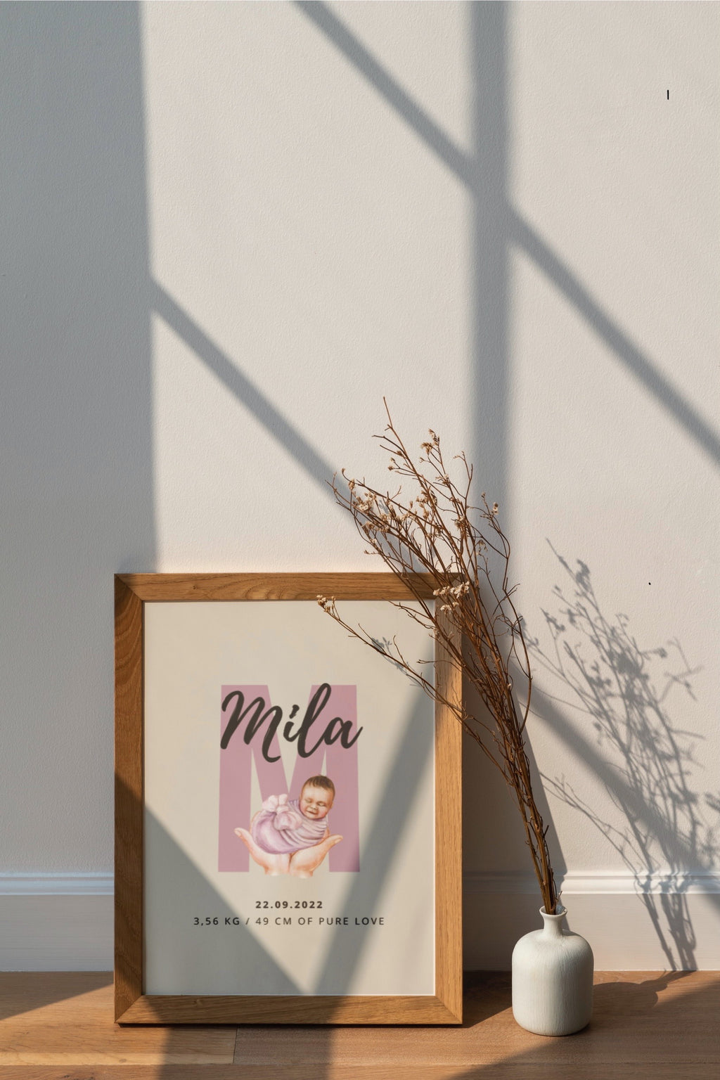 Framed photo of a baby with 'Mila' on a wall with a vase and branches.