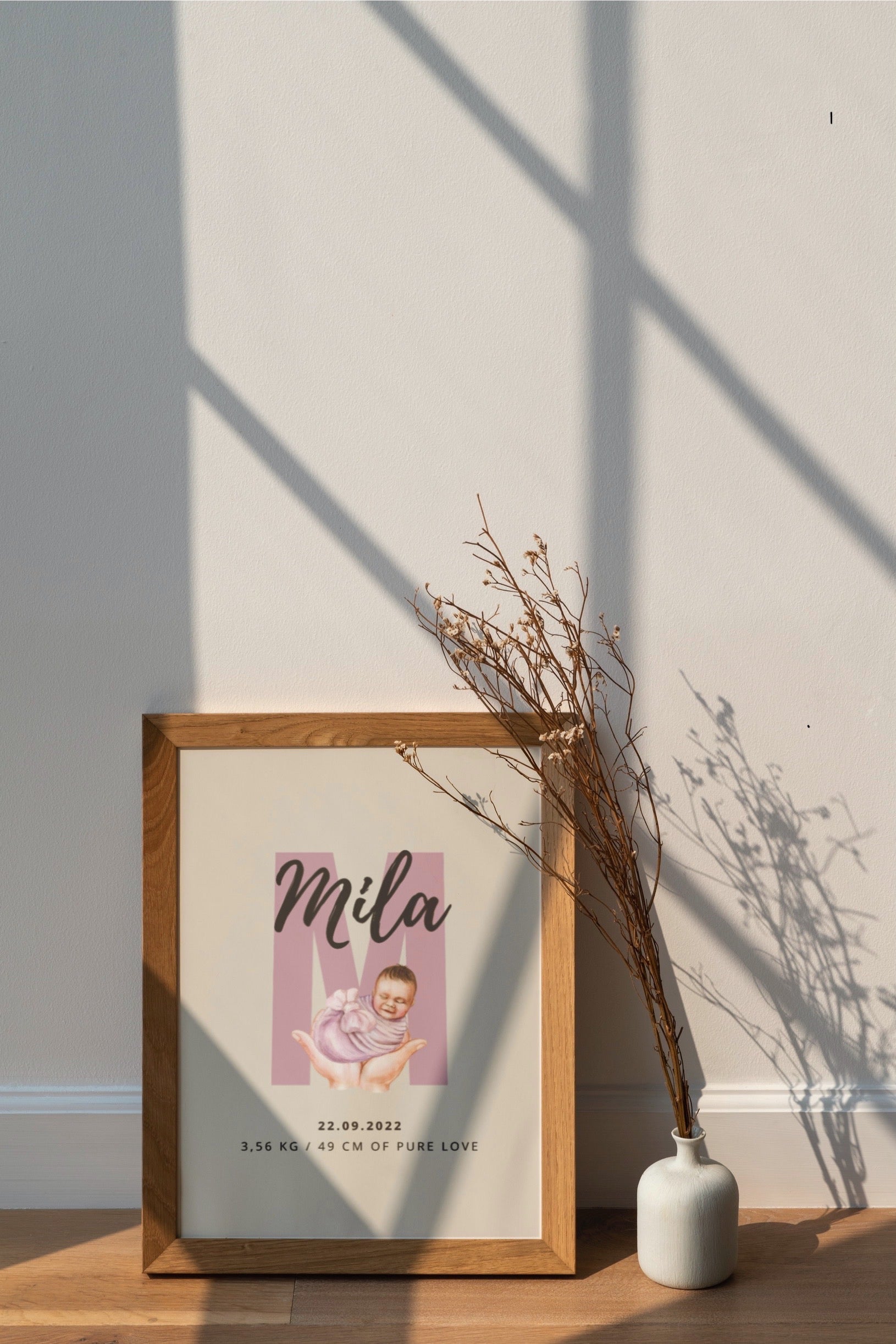 Framed photo of a baby with 'Mila' on a wall with a vase and branches.